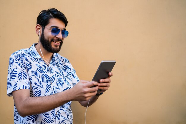 Porträt des jungen lateinischen Mannes, der sein digitales Tablett mit Kopfhörern gegen gelbe Wand verwendet. Technologie und Stadtkonzept.