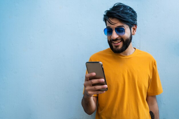 Porträt des jungen lateinischen Mannes, der sein Handy gegen den blauen Raum benutzt. Kommunikationskonzept.