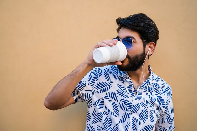 Porträt des jungen lateinischen Mannes, der Sommerkleidung trägt, eine Tasse Kaffee trinkt und Musik mit Kopfhörern gegen gelben Raum hört.