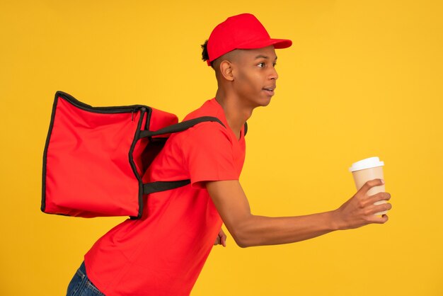 Porträt des jungen Lieferboten in roter Uniform, der mit einem Kaffee zum Mitnehmen läuft. Lieferservice-Konzept.