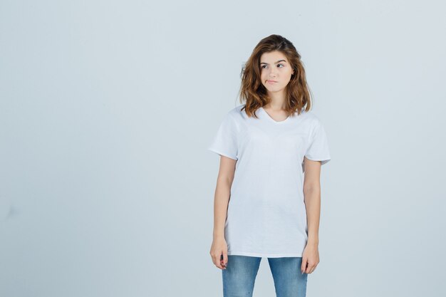 Porträt des jungen Mädchens, das beiseite schaut, gekrümmte Lippen im weißen T-Shirt und nachdenkliche Vorderansicht schaut