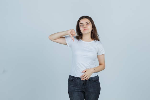 Porträt des jungen Mädchens, das Daumen unten in T-Shirt, Jeans zeigt und selbstbewusste Vorderansicht schaut