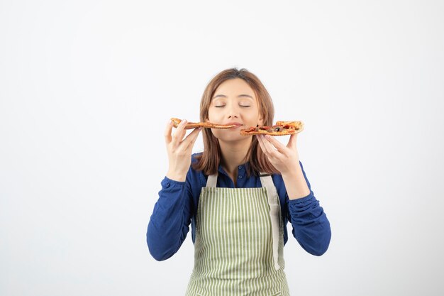 Porträt des jungen Mädchens in der Schürze, das Pizza auf Weiß isst