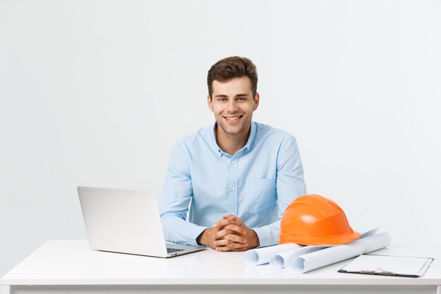 Porträt des jungen männlichen Innenarchitekten oder des Ingenieurs, der beim Sitzen auf seinem Bürotisch lächelt.