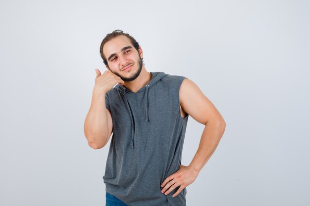 Porträt des jungen Mannes, der die Wange mit der Hand im ärmellosen Kapuzenpulli berührt und fröhliche Vorderansicht schaut