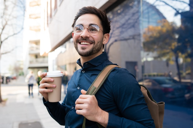 Porträt des jungen Mannes, der eine Tasse Kaffee hält, während draußen an der Straße geht. Stadt- und Lifestyle-Konzept.