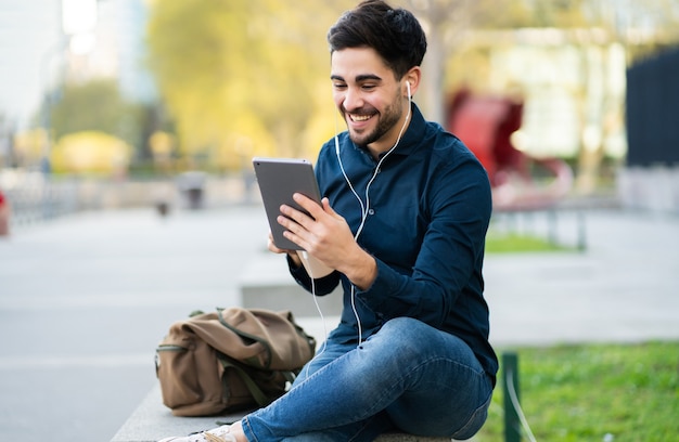 Porträt des jungen Mannes, der einen Videoanruf auf digitalem Tablett beim Sitzen auf Bank im Freien hat. Stadtkonzept.