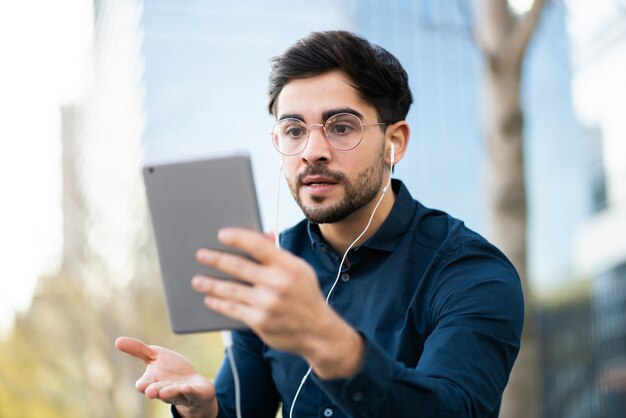 Porträt des jungen Mannes, der einen Videoanruf auf digitalem Tablett beim Stehen auf Bank im Freien hat