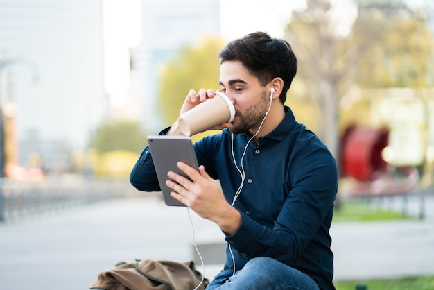 Porträt des jungen Mannes, der einen Videoanruf auf digitalem Tablett und Kaffeetrinken beim Sitzen auf Bank im Freien hat. Stadtkonzept.