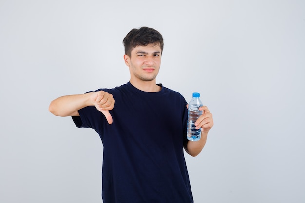 Porträt des jungen Mannes, der Flasche Wasser hält, Daumen unten im schwarzen T-Shirt zeigend und unzufriedene Vorderansicht schauend zeigt