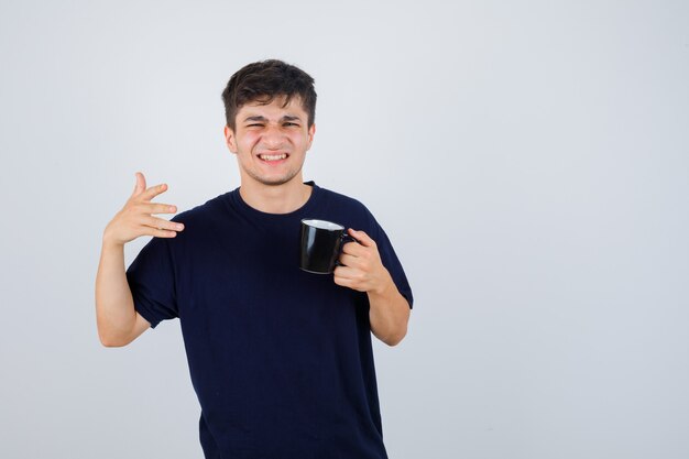 Porträt des jungen Mannes, der Tasse Tee hält, in schwarzem T-Shirt wegweisend und verwirrte Vorderansicht schauend zeigt