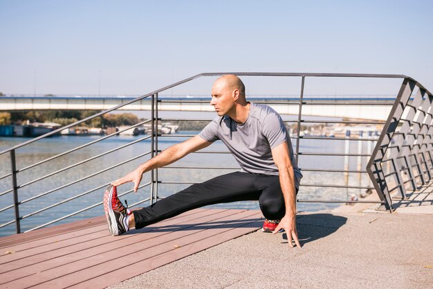 Porträt des jungen Mannes des Sitzes, der Bein auf Brücke nahe dem See ausdehnt