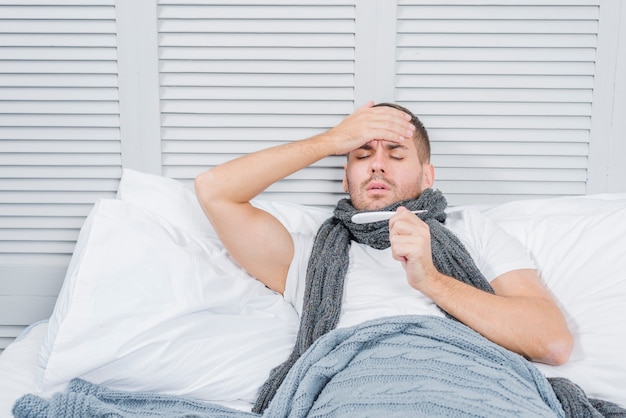 Porträt des jungen Mannes liegend auf dem Bett, das sein Fieber im Thermometer überprüft