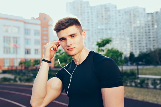 Porträt des jungen Mannes mit einem Sportkörper am Morgen auf Stadion. Er trägt ein schwarzes T-Shirt, legt einen Kopfhörer ans Ohr und sieht ernst aus.