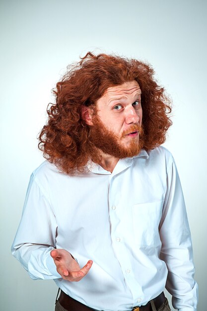 Porträt des jungen Mannes mit langen roten Haaren und mit schockiertem Gesichtsausdruck auf grauem Hintergrund