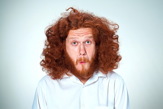 Porträt des jungen Mannes mit langen roten Haaren und mit schockiertem Gesichtsausdruck auf grauem Hintergrund