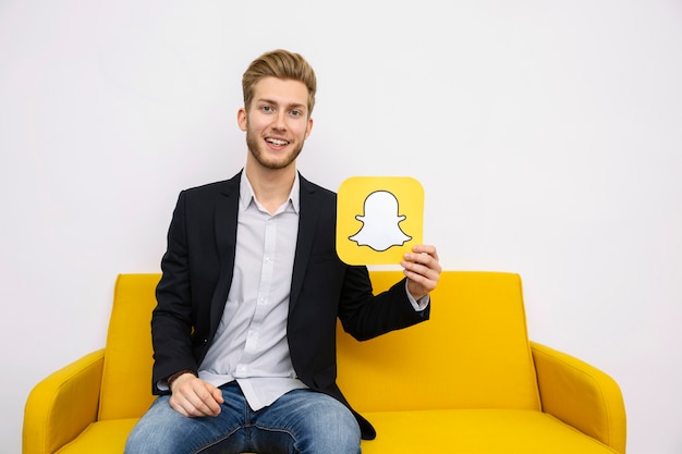 Kostenloses Foto porträt des jungen mannes sitzend auf dem gelben sofa, das snapchat ikone hält