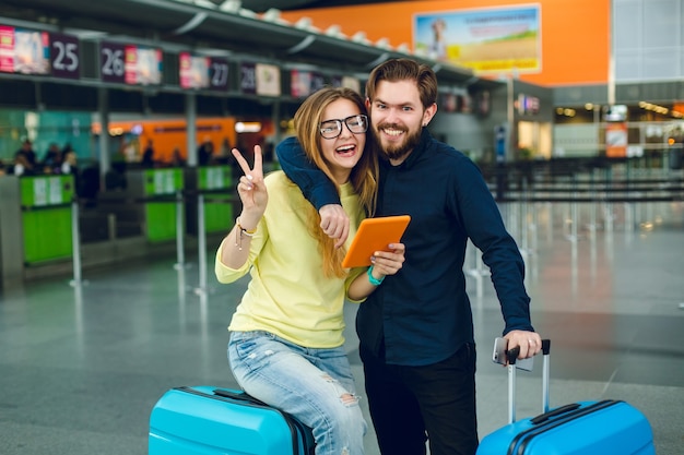 Porträt des jungen Paares, das im Flughafen umarmt. Sie hat lange Haare, einen gelben Pullover, Jeans und ein Tablet. Er hat ein schwarzes Hemd, eine Hose und einen Koffer in der Nähe. Sie lächeln in die Kamera.