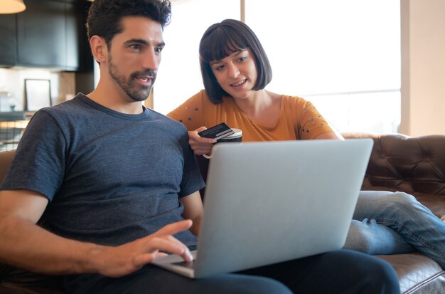 Porträt des jungen Paares, das online mit einer Kreditkarte und einem Laptop von zu Hause aus einkauft. E-Commerce-Konzept. Neuer normaler Lebensstil.