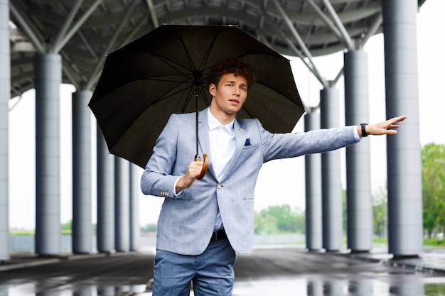 Porträt des jungen rothaarigen Geschäftsmannes, der das Auto fängt und Regenschirm hält