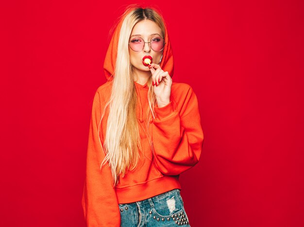 Porträt des jungen schönen bösen Hipster-Mädchens im trendigen roten Kapuzenpulli und im Ohrring in der Nase. Positives Modell leckt runde Kandiszucker