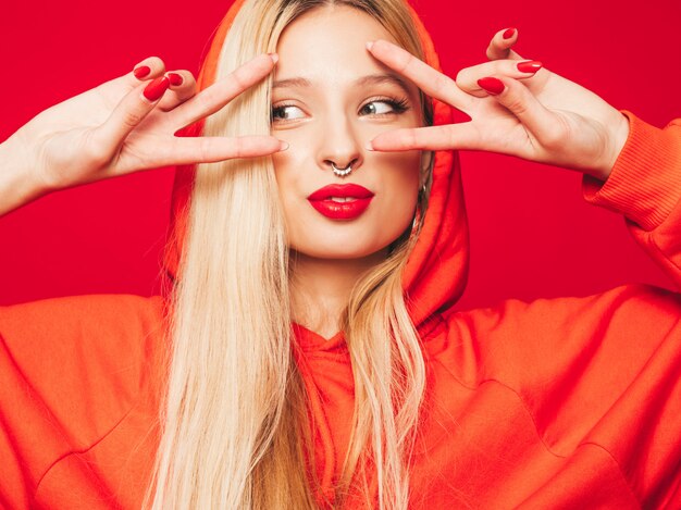 Porträt des jungen schönen bösen Hipster-Mädchens im trendigen roten Kapuzenpulli und im Ohrring in der Nase. Positives Modell zeigt Friedenszeichen