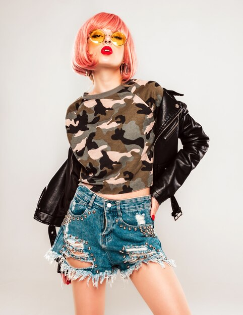 Porträt des jungen schönen Hipster bösen Mädchens in der schwarzen Lederjacke und im Ohrring in ihrer Nase. Sexy sorglose lächelnde Frau, die im Studio in der rosa Perücke nahe blauer Wand sitzt. Selbstbewusstes Modell in der Sonnenbrille