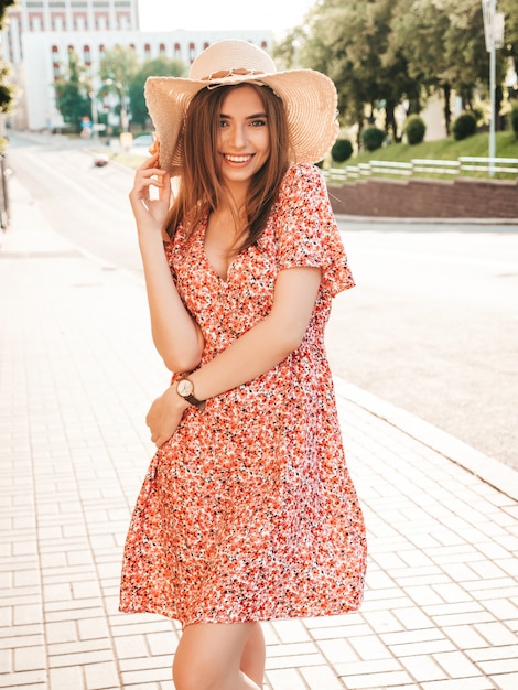 Porträt des jungen schönen lächelnden Hipster-Mädchens im trendigen Sommer-Sommerkleid. Sexy sorglose Frau, die auf dem Straßenhintergrund im Hut bei Sonnenuntergang aufwirft. Positives Modell im Freien