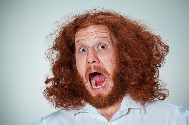 Porträt des jungen schreienden Mannes mit langen roten Haaren und mit schockiertem Gesichtsausdruck auf grauem Hintergrund
