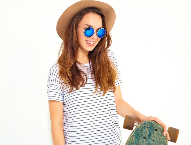 Porträt des jungen stilvollen Frauenmodells im zufälligen Sommer kleidet im braunen Hut, der mit longboard Schreibtisch aufwirft. Isoliert auf weiss