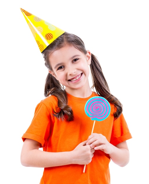 Porträt des kleinen niedlichen Mädchens im orangefarbenen T-Shirt und im Partyhut mit farbigen Süßigkeiten - lokalisiert auf Weiß