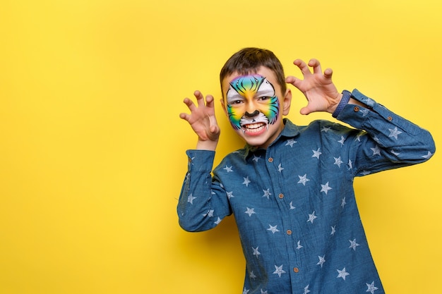 Porträt des kleinen süßen Jungen mit Faceart auf Geburtstagsfeier, süßer bunter Tiger einzeln auf gelber Wand.