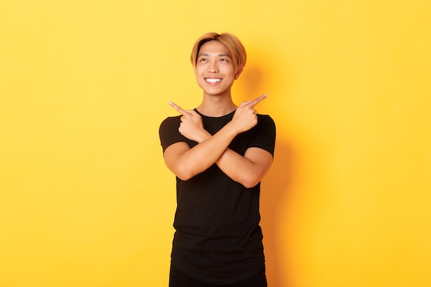 Porträt des lächelnden erfreuten asiatischen Kerls, der nach links schaut und Entscheidung trifft, seitwärts zeigt, gelbe Wand