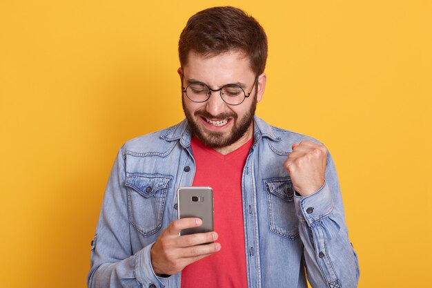Porträt des lächelnden glücklichen Mannes, der Jeansjacke und rotes Hemd trägt, Faust ballt und Smartphone in Händen hält
