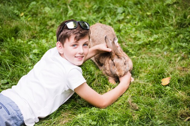 Porträt des lächelnden Jungen liegend auf dem grünen Gras, das um seinem Kaninchen sich kümmert