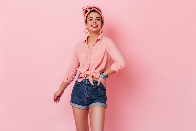 Porträt des lächelnden Mädchens im Pin-up-Stil. Frau mit rosa Haarbandage gekleidet in Hemd und Shorts, die auf isoliertem Raum aufwerfen.