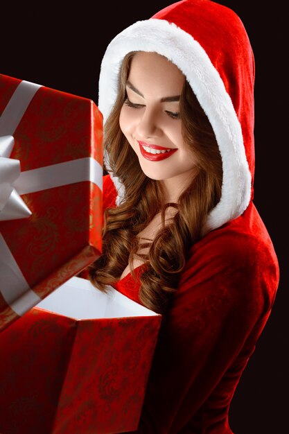 Porträt des lächelnden Mädchens im roten Anzug, das ein Geschenk für Neujahr öffnet
