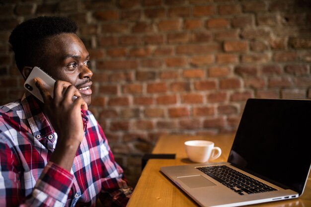 Porträt des lächelnden Mannes, der auf Handy spricht, während er in einem Café mit einem Laptop sitzt
