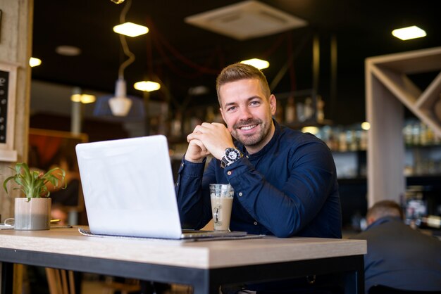 Porträt des lächelnden Mannes, der in einer Café-Bar mit seinem Laptop-Computer sitzt