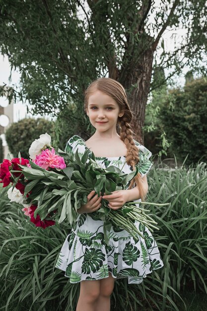 Porträt des lächelnden schönen Teenager-Mädchens mit Blumenstrauß der Pfingstrosen gegen grünes Gras am Sommerpark. Kindermode-Konzept.