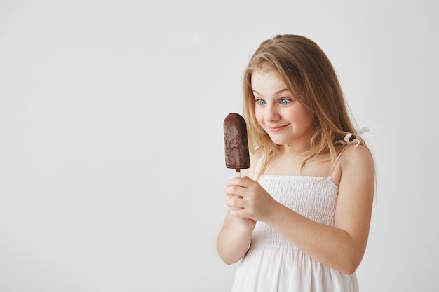 Porträt des lustigen blonden kleinen Haares im weißen Kleid, das Eis in ihren Händen mit glücklichem und aufgeregtem Blick betrachtet und es essen wird.