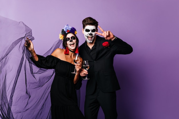 Porträt des lustigen Kerls und des Mädchens mit gemalten Gesichtern, die Spaß mit Gläsern Wein auf Halloween-Party haben.