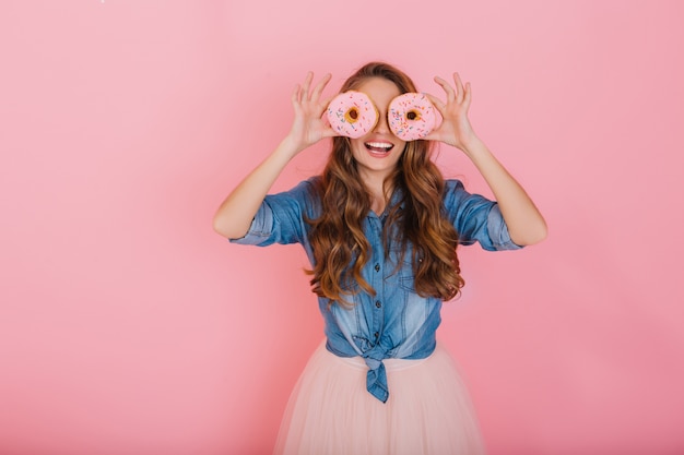 Porträt des mädchens mit dem langen haar, das rosa donuts als brille und lachen lokalisiert auf rosafarbenem hintergrund hält. entzückende lächelnde junge brünette junge frau, die spaß mit donuts nach dem teetrinken hat