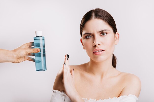 Porträt des Mädchens mit unzufriedenem Gesichtsausdruck auf weißer Wand mit Mizellenwasser. Frau ohne Make-up gegen die Verwendung von Kosmetika.