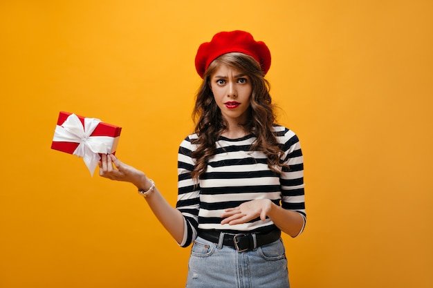 Porträt des Mädchens sieht unglücklich aus und hält Geschenkbox. Moderne junge Frau im roten Baskenmützen- und Jeansrock mit dem schwarzen Gürtel, der aufwirft.