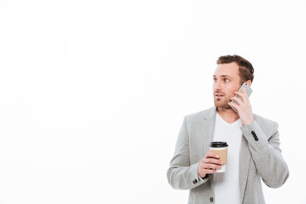 Porträt des männlichen Büroangestellten Kaffee zum Mitnehmen beim Haben des angenehmen beweglichen Gespräches auf Mobiltelefon und beiseite schauen, über weißer Wand trinkend