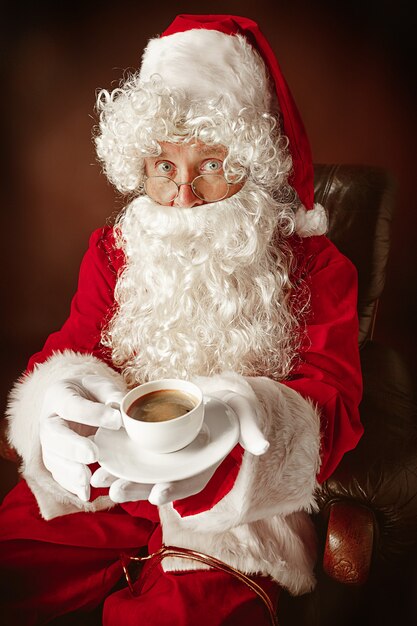 Porträt des Mannes im Weihnachtsmann-Kostüm - mit einem luxuriösen weißen Bart, der Weihnachtsmann-Mütze und einem roten Kostüm am roten Studiohintergrund, der in einem Stuhl mit Tasse Kaffee sitzt