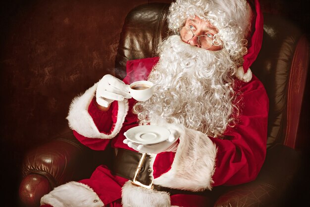 Porträt des Mannes im Weihnachtsmann-Kostüm - mit einem luxuriösen weißen Bart, der Weihnachtsmann-Mütze und einem roten Kostüm am roten Studiohintergrund, der in einem Stuhl mit Tasse Kaffee sitzt