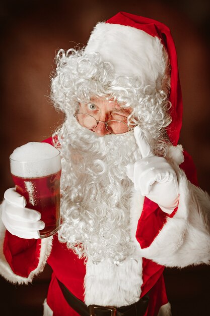 Porträt des Mannes im Weihnachtsmann-Kostüm - mit einem luxuriösen weißen Bart, der Weihnachtsmann-Mütze und einem roten Kostüm am roten Studiohintergrund mit Bier