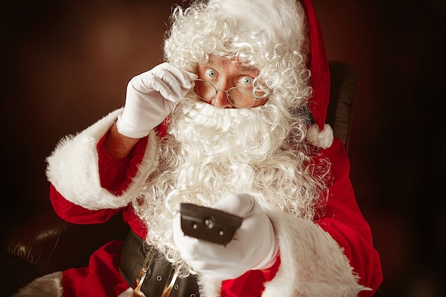 Porträt des Mannes im Weihnachtsmann mit einem luxuriösen weißen Bart, der Weihnachtsmannmütze und einem roten Kostüm am roten Studiohintergrund